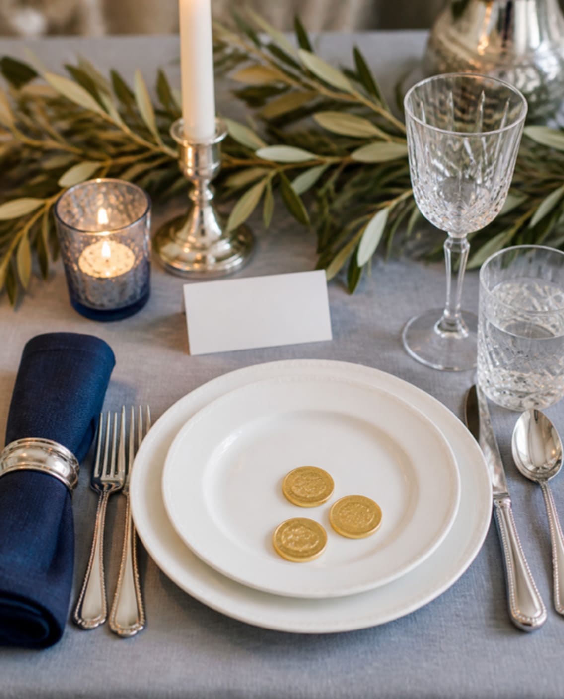 Mood board: Mood board of one Hanukkah place setting: three gold coins on a white plate and a navy napkin in a silver ring.